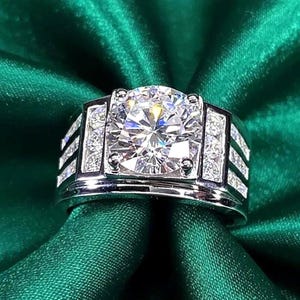 May include: A silver ring featuring a large, clear, round-cut gemstone at its centre. Smaller, clear gemstones are set on the sides. The ring is displayed on green, textured fabric.