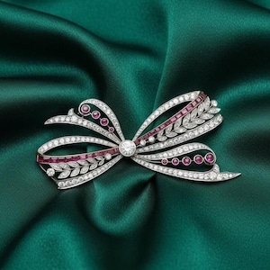 Op de afbeelding: Een strikvormige broche met een centrale diamant en accenten van rode edelstenen en kleinere diamanten. De broche is gezet in een zilverkleurig metaal en ligt op een donkergroene satijnen stof.