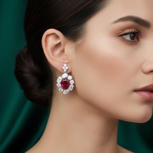 Puede incluir: Primer plano del rostro de una mujer, destacando un deslumbrante pendiente. El pendiente presenta una gema central ovalada de color rojo intenso, rodeada de gemas redondas más pequeñas y transparentes. El engaste es plateado.