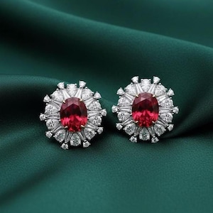 May include: Two stud earrings with oval, deep red gemstones, encircled by smaller, clear stones. The earrings are set against a dark green backdrop, accentuating their brilliance and design. A classic jewellery piece.