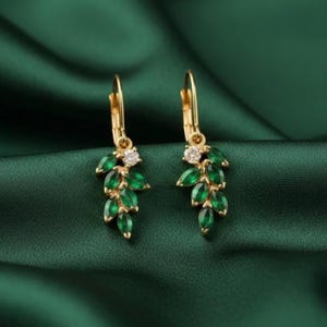 May include: A pair of gold-colored leaf-shaped earrings with green and clear gemstones. The earrings have a lever back closure and are set against a dark green fabric background.
