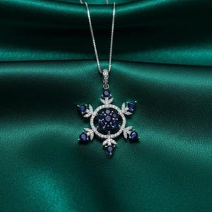 Könnte beinhalten: Eine silberfarbene Schneeflocken-Anhänger-Halskette mit einer filigranen Kette. Der Anhänger hat ein Schneeflocken-Design mit einem zentralen Cluster aus dunkelblauen Edelsteinen, umgeben von einem Ring aus klaren Steinen und sechs äußeren Spitzen, jede mit einem blauen Edelstein. Die Halskette ist vor einem grünen Satinhintergrund platziert.