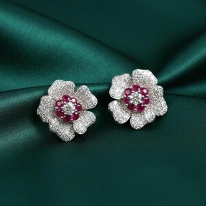 Puede incluir: Un par de pendientes de botón con forma de flor. Cada pendiente presenta un grupo de pequeñas piedras preciosas rojas redondas en el centro, rodeado por un anillo de piedras claras y brillantes. Los pétalos también están cubiertos de piedras claras.