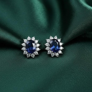 Puede incluir: Un par de elegantes pendientes de botón con piedras preciosas ovales de color azul oscuro, rodeadas por piedras más pequeñas y claras. Los pendientes se presentan sobre un fondo verde oscuro y sedoso, creando una estética lujosa.