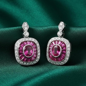 May include: A pair of elegant earrings featuring a central oval ruby surrounded by smaller rubies and a border of sparkling white gemstones. The earrings have a rectangular shape with a diamond-shaped top and are set against a dark green background.
