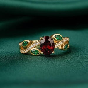 May include: A gold ring with a large oval, deep red gemstone as the focal point. The ring is adorned with smaller green gemstones and tiny white stones. The band has a leaf-like design. The ring is set against a dark green background.