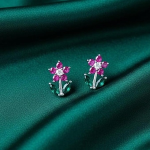 Może przedstawiać: Para kolczyków w kształcie kwiatów. Każdy kolczyk ma różowe kamienie szlachetne tworzące płatki, centralny biały kamień szlachetny i zielone listki z kamieni szlachetnych. Kolczyki są umieszczone na ciemnozielonym satynowym tle.