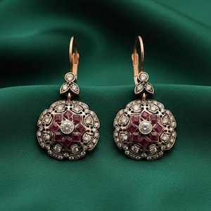 May include: A pair of gold-coloured earrings with a floral design. Each earring has a central round clear stone, surrounded by square red stones and smaller clear stones. The earrings hang from a gold-coloured hook.