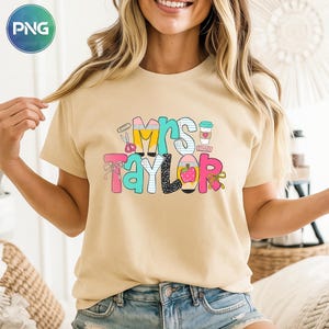 May include: A beige t-shirt featuring the text "Mrs. Taylor" in a vibrant, block letter design. The graphic incorporates school-related imagery such as scissors, pencils, an apple, and a coffee cup. The shirt is a casual, short-sleeved style.