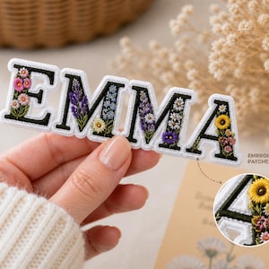 May include: Embroidered patch spelling "EMMA" with black letters and white borders. Each letter is filled with colourful floral embroidery. The patch is held by a person wearing a white sweater. The text "EMBROIDERED PATCHES" is also visible.