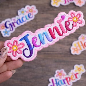 Puede incluir: Parche bordado con el nombre "Jennie" en letras de colores. El parche está delineado en rosa con un diseño floral y un fondo blanco. Se ven parches adicionales con nombres.