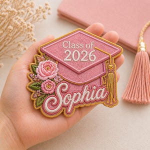 May include: A pink embroidered graduation patch featuring a mortarboard, flowers, and the text "Class of 2026" and "Sophia". The patch has a gold border and a gold tassel.