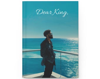 May include: A hardcover journal with a teal cover featuring the text "Dear King," in elegant script. The image on the cover depicts a person overlooking a bright blue ocean.