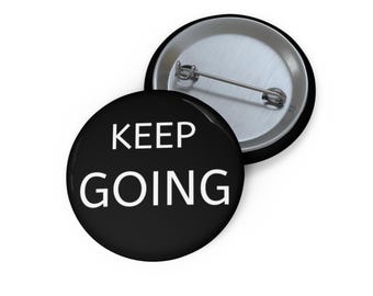 May include: A black button with the words "KEEP GOING" in white, with a silver pin back. The button is round and has a shiny, reflective surface. Another button is partially visible in the background.