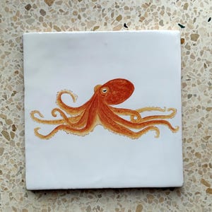 May include: A square, white ceramic tile with a watercolour illustration of an orange octopus. The octopus has eight tentacles and is centred on the tile. The tile has a smooth, glossy finish.