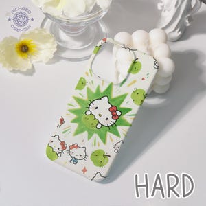 Puede incluir: Funda de tel&eacute;fono blanca con un dise&ntilde;o de Hello Kitty, manzanas verdes y un patr&oacute;n de explosi&oacute;n verde. La palabra "HARD" est&aacute; impresa en negro. La funda tiene una parte superior curvada para la c&aacute;mara.