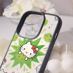 Puede incluir: Funda de tel&eacute;fono con un dise&ntilde;o de Hello Kitty blanco con un lazo rojo, sosteniendo un objeto verde, sobre un fondo blanco con acentos verdes. La funda tiene un borde gris oscuro.