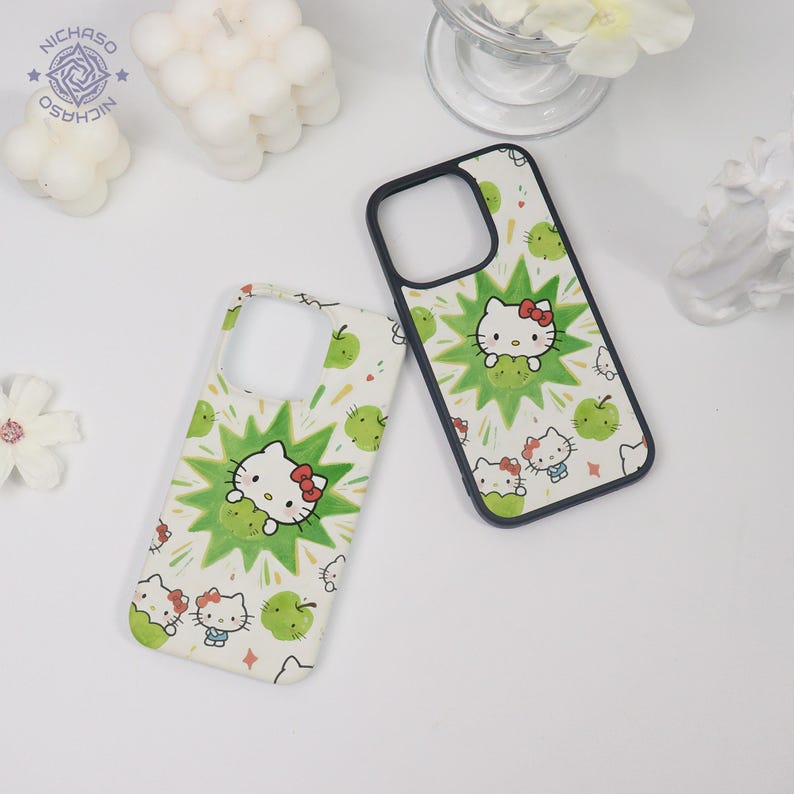 Puede incluir: Dos fundas para tel&eacute;fono sobre un fondo blanco. Las fundas presentan un patr&oacute;n repetido de manzanas verdes y personajes de Hello Kitty. Cada funda tiene un borde negro y un recorte para la c&aacute;mara. Una funda tiene un borde blanco.