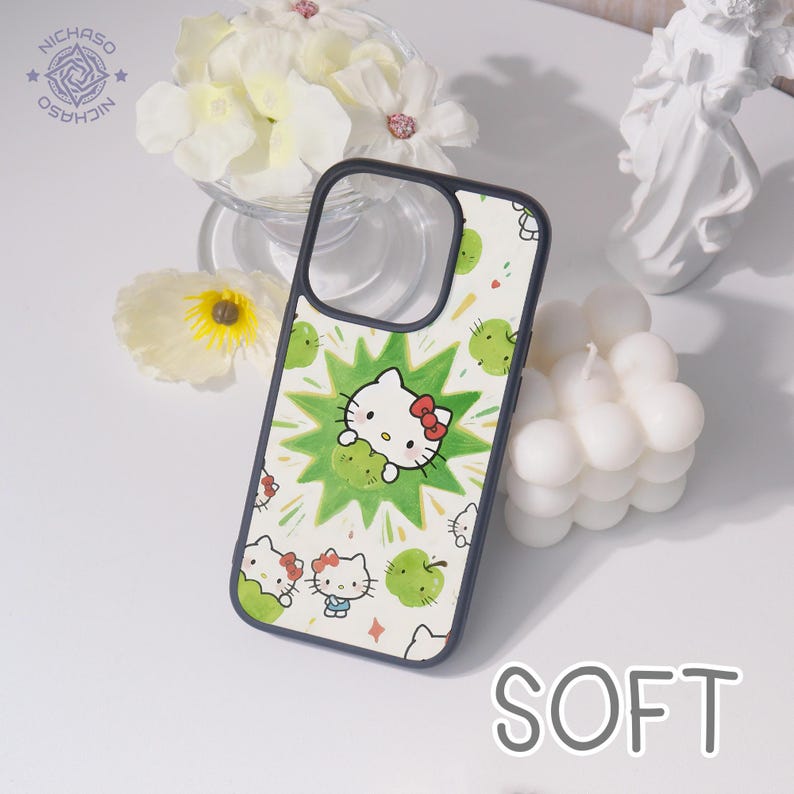 Puede incluir: Funda de tel&eacute;fono con fondo blanco adornada con manzanas verdes y motivos de Hello Kitty. El dise&ntilde;o central muestra a Hello Kitty con un lazo rojo. La funda tiene un borde negro. La palabra "SOFT" es visible.