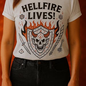 Op de afbeelding: Wit t-shirt met de tekst "HELLFIRE LIVES!" boven een schildontwerp met een schedel met hoorns, omringd door vlammen, zwaarden en geometrische vormen. Het shirt wordt gedragen met een zwarte jeans.