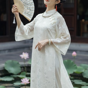 May include: A cream-colored dress featuring a high collar, three decorative white buttons on the upper chest, and wide, three-quarter length sleeves. The fabric is adorned with a tonal floral pattern. A folding hand fan with a white tassel is held.