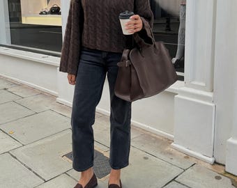 Op de afbeelding: Een vrouw draagt een bruine kabelgebreide trui, een donkere spijkerbroek en bruine loafers. Ze draagt een bruine leren tote bag en een wegwerp koffiebeker. Ze draagt ook een zonnebril. De outfit is gestyled voor een casual, alledaagse look.