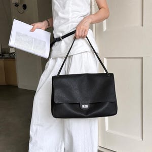 以下が含まれることがあります： 長方形のシルエット、大きなフロントフラップ、シルバーカラーの金属製ターンロック式留め具を備えた黒いレザーのショルダーバッグ。バッグの上部には2本の細いショルダーストラップが付いており、全体に質感のあるグレイン仕上げが施されています。