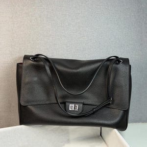 Pode incluir: Uma bolsa de ombro preta com textura granulada, apresentando uma aba frontal grande, alças de ombro finas e ajustáveis, e um fecho giratório de metal prateado de formato retangular centralizado na aba. A bolsa possui um formato estruturado.
