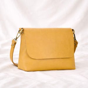 Könnte beinhalten: Senfgelbe Leder-Umhängetasche mit Klappenverschluss. Die Tasche hat einen langen, verstellbaren Riemen und goldfarbene Beschläge. Die Tasche ist rechteckig und liegt auf einer weißen Oberfläche.