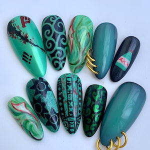 May include: A collection of ten almond-shaped artificial nails. The nails feature various designs, including swirls, geometric patterns, and glitter, in shades of green, black, and red. Some nails have gold rings attached. The overall aesthetic is artistic and modern.
