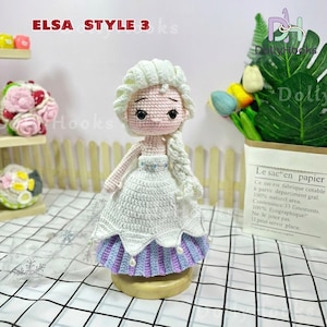 May include: A handmade crochet doll in the style of Elsa from Frozen. The doll has a white dress with a blue and purple skirt, a white braided hairstyle, and the text "ELSA STYLE 3 ks". The doll is on a wooden base.