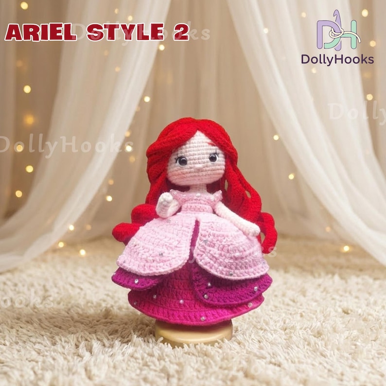 May include: A handmade crochet doll in the style of Ariel, featuring red hair and a pink and purple dress. The doll has a detailed face and is displayed on a gold-colored base. The text "ARIEL STYLE 2s" is visible.