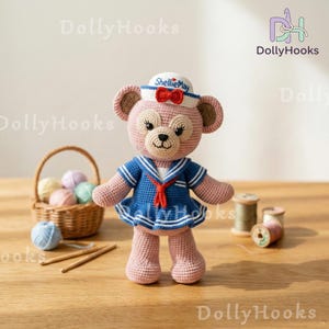 May include: A handmade crochet bear doll wearing a sailor outfit. The pink bear has a blue sailor dress with white stripes and a red bow. The bear is wearing a white sailor hat with the text "ShellieMay". A basket of yarn and crochet hooks are in the background.