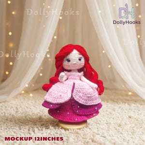 May include: A handmade crochet doll with red hair and a pink and purple dress. The doll is approximately 12 inches tall. The doll has a white face with black eyes and is standing on a gold base. The text "DollyHooks" is visible.