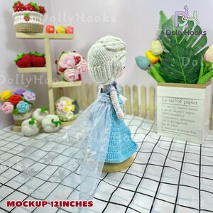 May include: A handmade crochet doll, approximately 12 inches tall, dressed in a blue gown with a sheer, sparkling cape. The doll has a white crocheted hairstyle. The background includes other crochet items like flowers and strawberries.