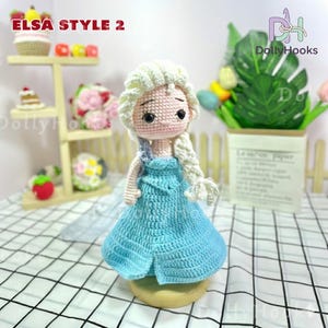 May include: A handmade crochet doll in the style of Elsa from Frozen. The doll has a light blue dress, blonde hair in a braid, and is standing on a wooden base. The text "ELSA STYLE 2" is visible in the upper left corner.