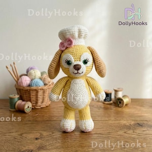 May include: A handmade crochet dog wearing a chef's hat and a pink bow. The dog is yellow with white accents and green eyes. The background includes a basket of yarn, wooden spools, and the DollyHooks logo.