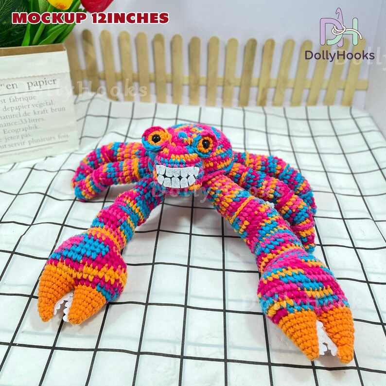 K&ouml;nnte beinhalten: Ein farbenfrohes H&auml;kelkrabben-Spielzeug, ca. 30 cm lang, mit einem l&auml;chelnden Gesicht und gro&szlig;en Scheren. Die Krabbe besteht aus rosa, blauer und orangefarbener Wolle. Der Text "MOCKUP 12INCHES" ist oben links sichtbar.