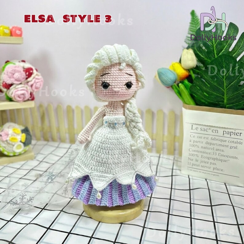 May include: A handmade crochet doll in the style of Elsa from Frozen. The doll has a white dress with a blue and purple skirt, a blonde braid, and the text "ELSA STYLE 3 ks". The doll is on a wooden base.