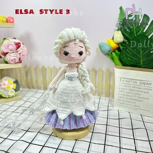 May include: A handmade crochet doll in the style of Elsa from Frozen. The doll has a white dress with blue and purple accents, a blonde braid, and the text "ELSA STYLE 3 ks". The doll is on a wooden base.