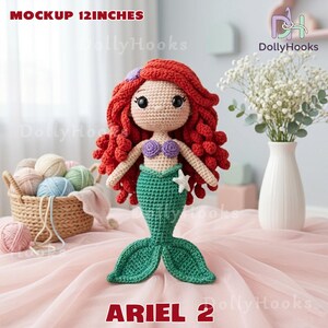 Puede incluir: Una mu&ntilde;eca de sirena de ganchillo, de aproximadamente 30 cm de altura, con cabello rojo, un top de concha p&uacute;rpura y una cola verde. La mu&ntilde;eca tiene una estrella de mar blanca y el texto "ARIEL 2". La mu&ntilde;eca se exhibe sobre una superficie rosa.