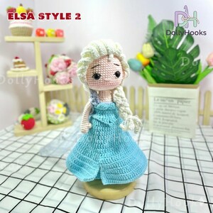 May include: Crocheted doll in the style of Elsa from Frozen, featuring a light blue dress and blonde braided hair. The doll has a pink face and is displayed on a wooden base. The text "ELSA STYLE 2" is visible in the image.