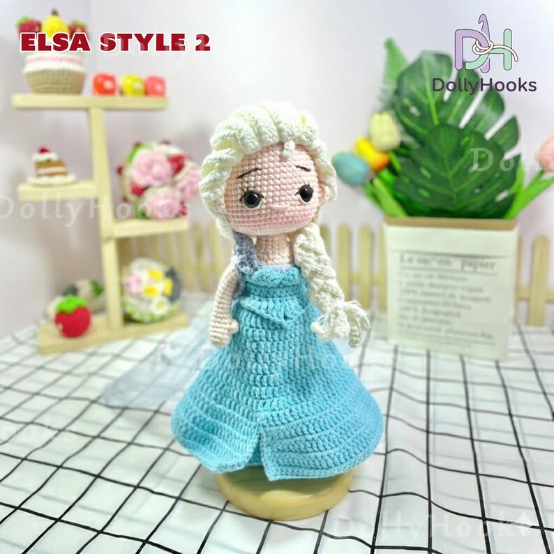 May include: Crocheted doll in the style of Elsa from Frozen, featuring a light blue dress and blonde braided hair. The doll has a pink face and is displayed on a wooden base. The text "ELSA STYLE 2" is visible in the image.