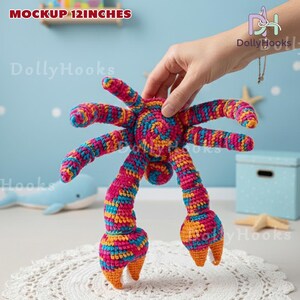 K&ouml;nnte beinhalten: Ein farbenfrohes, handgefertigtes H&auml;kelkrabben-Spielzeug, ca. 30 cm gro&szlig;. Die Krabbe hat eine Mischung aus rosa, blauem und gelbem Garn mit orangefarbenen Akzenten an ihren Scheren. Das Spielzeug wird von einer Hand gehalten, die seine detaillierte Handwerkskunst zeigt.