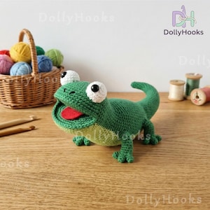 May include: A handmade green crochet lizard with a red tongue and large white eyes. The lizard is on a wooden surface, with a basket of colorful yarn balls and crochet hooks in the background. The lizard is a whimsical, handcrafted item.