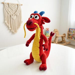 May include: A handmade red and yellow crochet dragon toy with blue horns and yellow whiskers. The dragon has large, expressive eyes and is posed on a white surface. The toy is likely intended for children and is part of a collection of handmade toys.