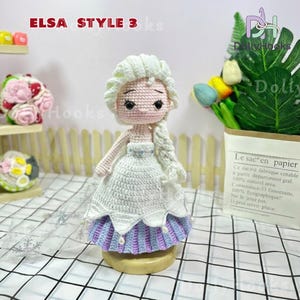 May include: Crocheted doll in the style of Elsa from Frozen, with a white dress, blue and purple skirt, and blonde braid. The doll is on a wooden base. Text at the top reads "ELSA STYLE 3 ks".