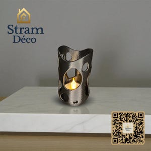 May include: A dark gray metal candle holder with an irregular, open design. The holder has several oval cutouts and contains a flickering LED candle. The product is displayed on a white surface. The brand name "Stram Déco" is visible in the image.