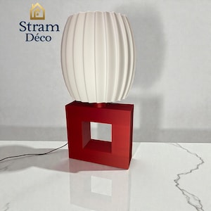 Puede incluir: Una lámpara de mesa moderna con una pantalla blanca y acanalada sobre una base cuadrada roja vibrante con un recorte central. La lámpara está conectada a un cable de alimentación. El texto "Stram Déco" es visible.
