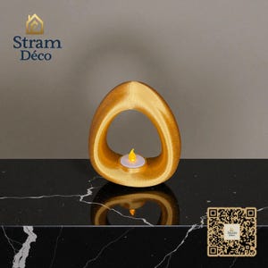 May include: A gold-colored, abstract candle holder with an oval shape and a small, lit LED candle inside. The candle holder is on a black surface. The text "Stram Déco" is in the upper left corner.